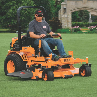 Man operating an orange SCAG lawn mower on a grassy area with a building and trees in the background.