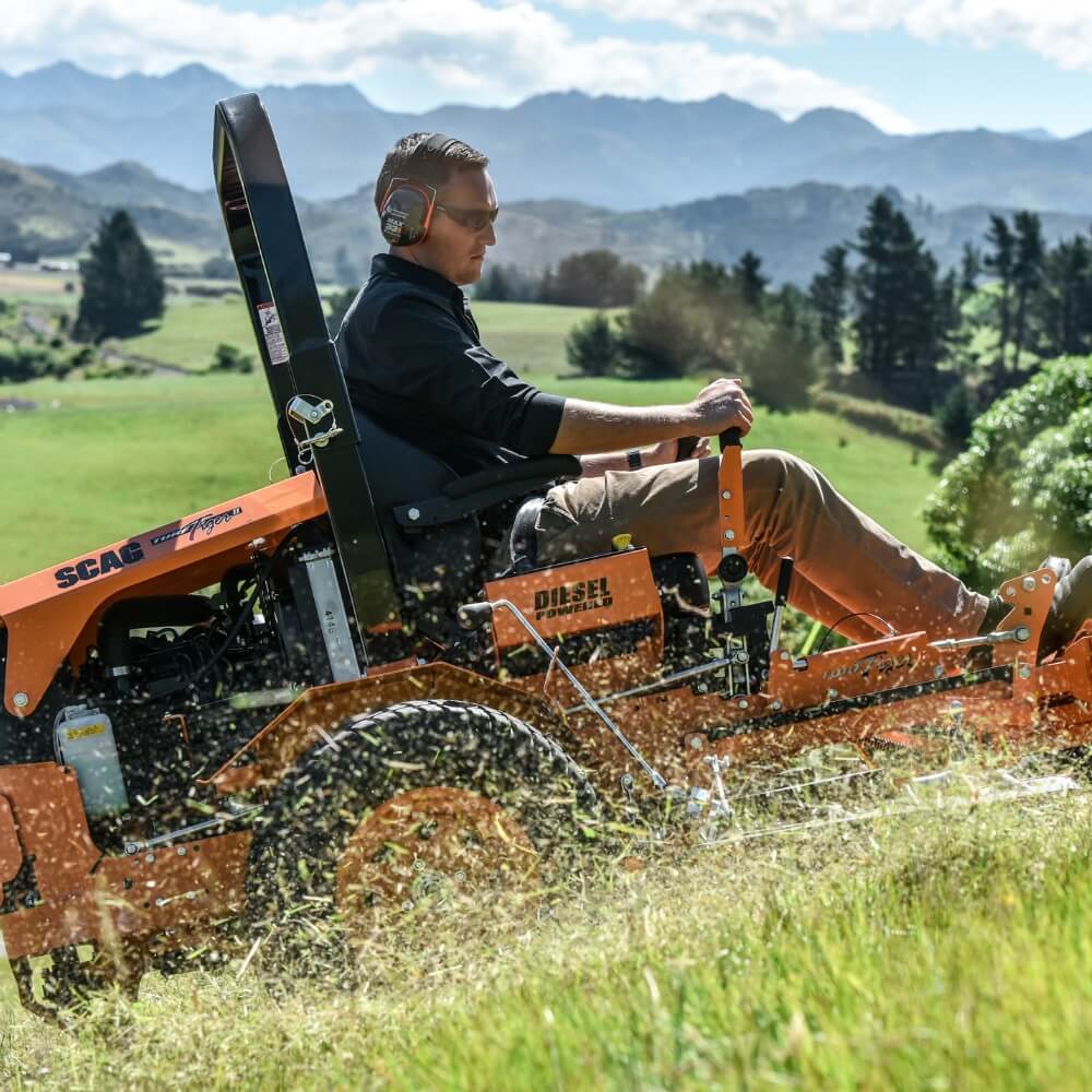 Person operating a SCAG mower in a grassy field in New Zealand with mountains in the background